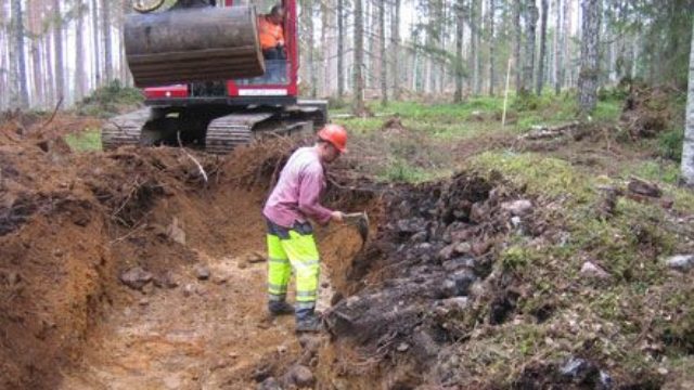 Arkeolog gräver ut ett röjningsröse i skogsmark. Grävmaskin i bakgrunden.