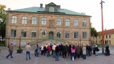 Ett stort tegelhus med grönt tak. Framför huset står det en stor folkmassa.