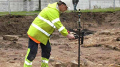 En man står i en jordgrop med ett verktyg. Bakom honom syns höghus och en sjö.