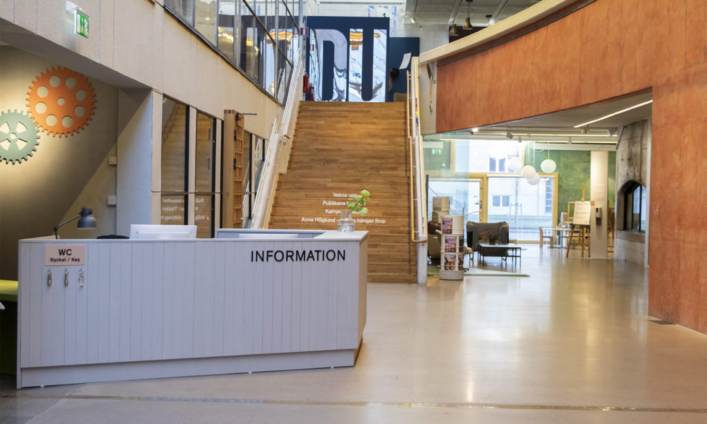 Grått träbord med svart bokstäver som formar ordet information. Bordet står på golvet och i bakgrunden syns en trappa som leder upp till övervåningen.