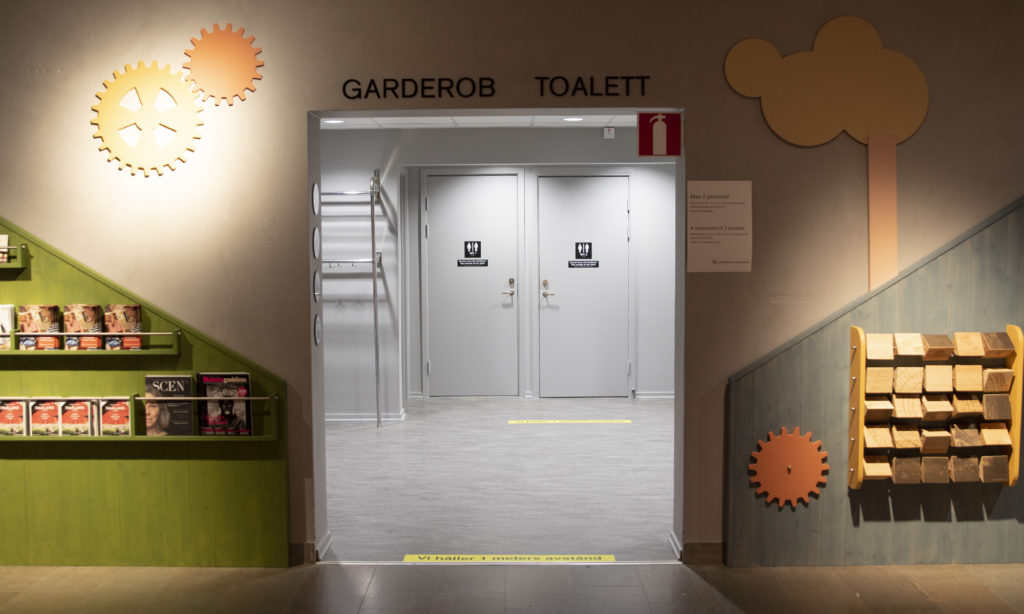 Ingång till museets kapprum. Till vänster finns ett grönt broschyrställ på väggen och till höger ett träfspel. Kapprummet är målat grått och mitt i kapprummet finns två dörrar till toaletter.