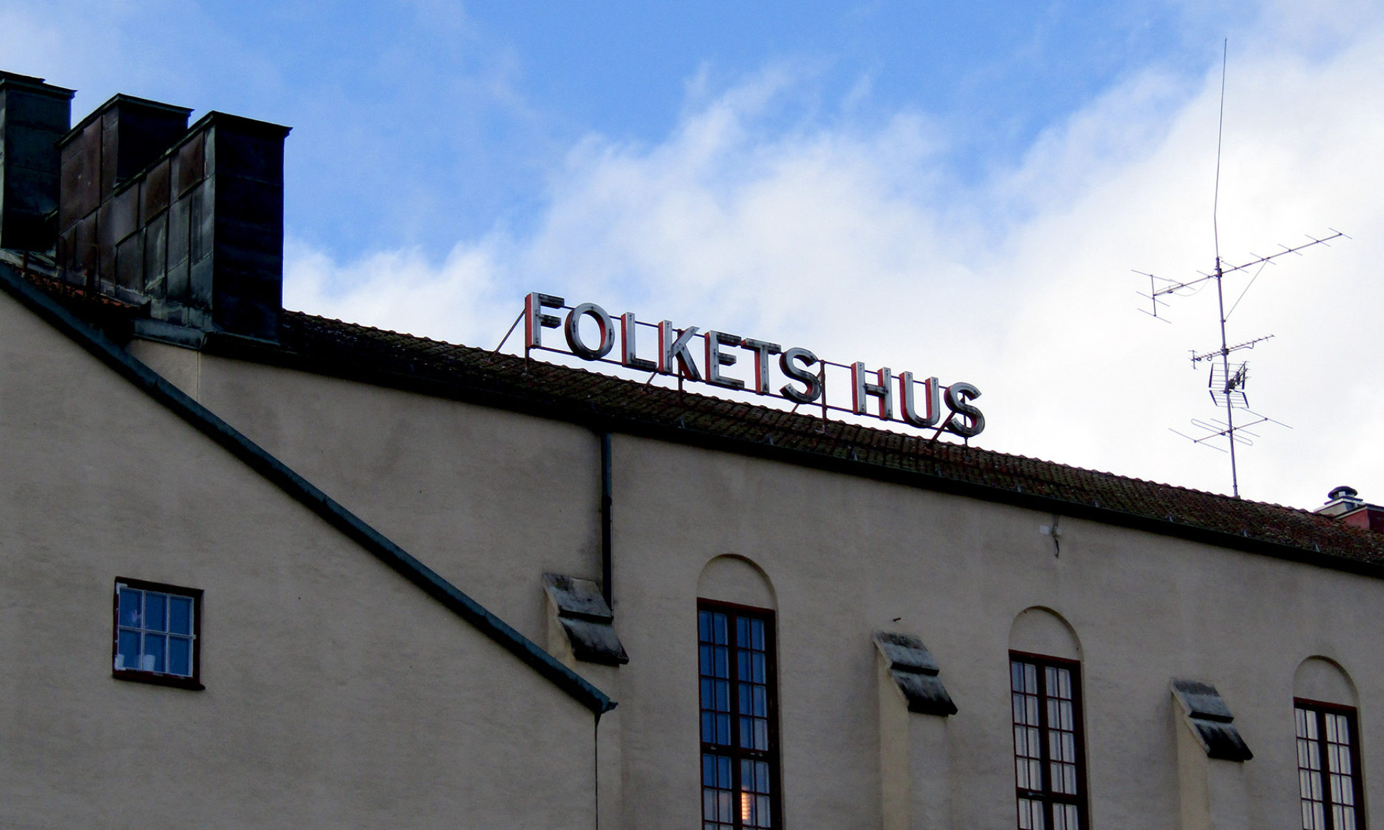 En byggnad mot blå himmel med moln. På taket står det Folkets hus med stora bokstäver.